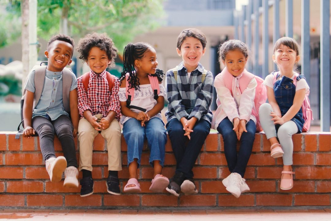Portrait Of Smiling Multi-Cultural Elementary School Pupils Sitting On Wall Outdoors At School